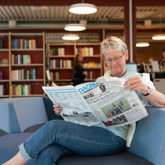 Kvinde, der læser en avis i en af de bløde sofagrupper i biblioteket. Bag hende ses reoler med bøger, samt selvbetjeningsautomaterne.