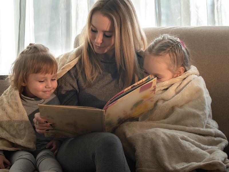 En kvinde sidder i en sofa med to børn. De deler et tæppe og læser i en bog sammen.