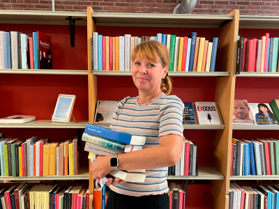 Pia Brun Jepsen, der holder en stak bøger. Hun står foran en bogreol ude i biblioteket.