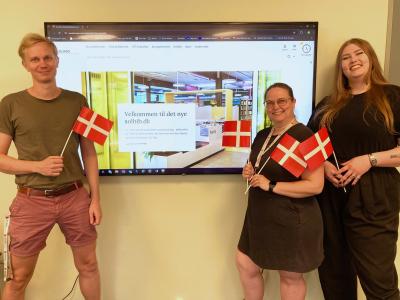 Bibliotekets systemadministratorgruppe (Rasmus, Lisbeth og Fie) foran en skærm med den nye hjemmeside på. De holder alle tre små flag og smiler.