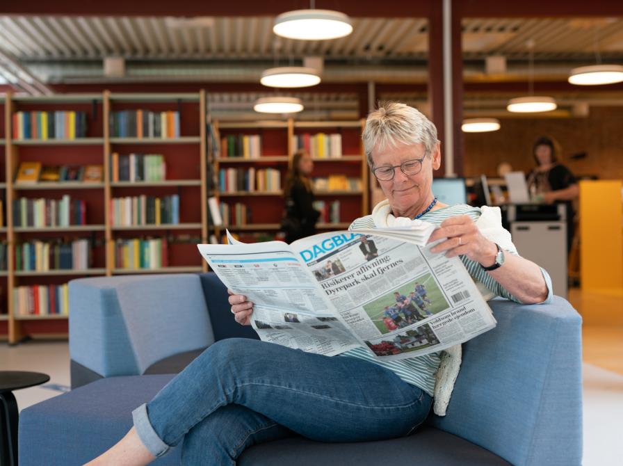 Kvinde, der læser en avis i en af de bløde sofagrupper i biblioteket. Bag hende ses reoler med bøger, samt selvbetjeningsautomaterne.